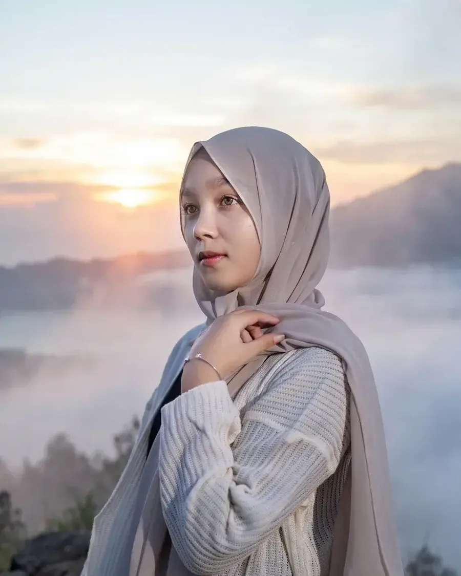 A woman in a beige hijab and a cream knit at a misty mountain sunrise